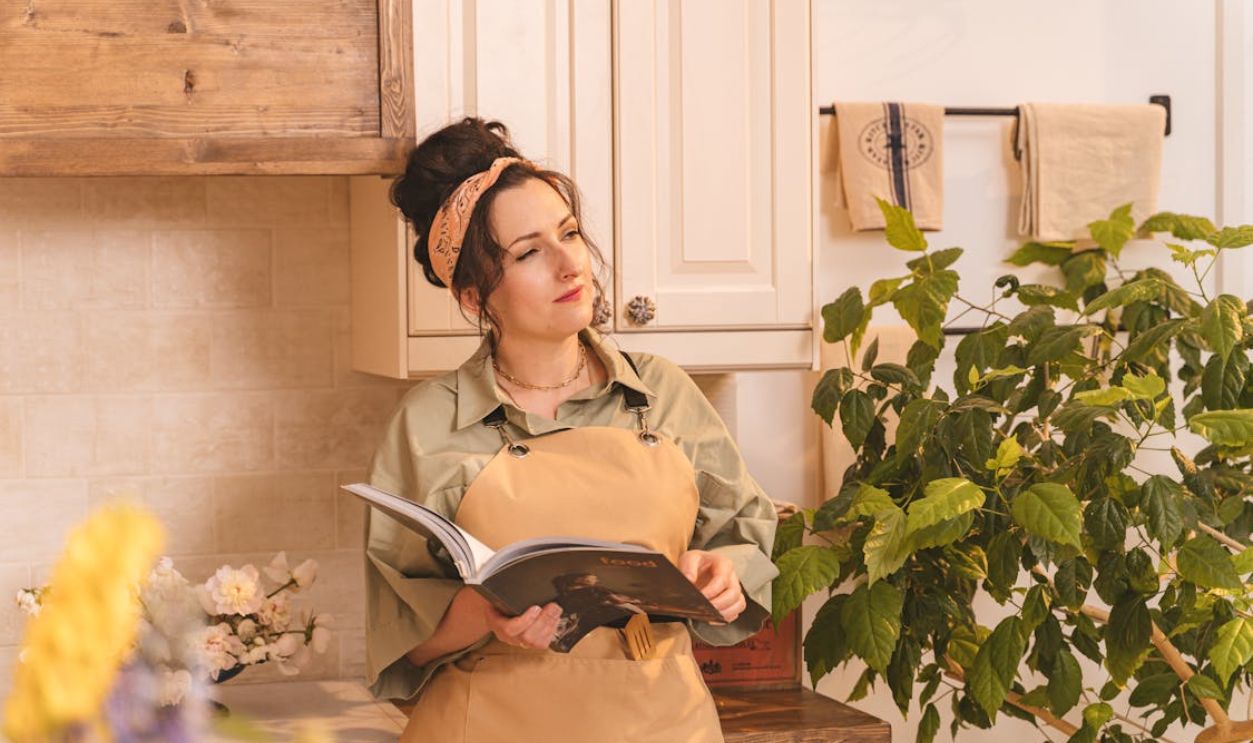 Woman in Brown Apron and Bandana Standing in the Kitchen Holding a Magazine