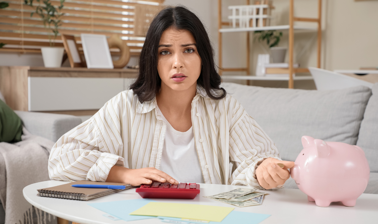 Tired young woman with piggy bank and calculator doing taxes at home.