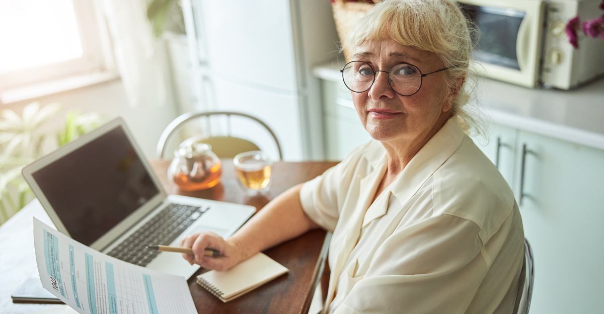 Nice elderly lady in glasses holding individual income tax return form and smiling while sitting at the table with laptop