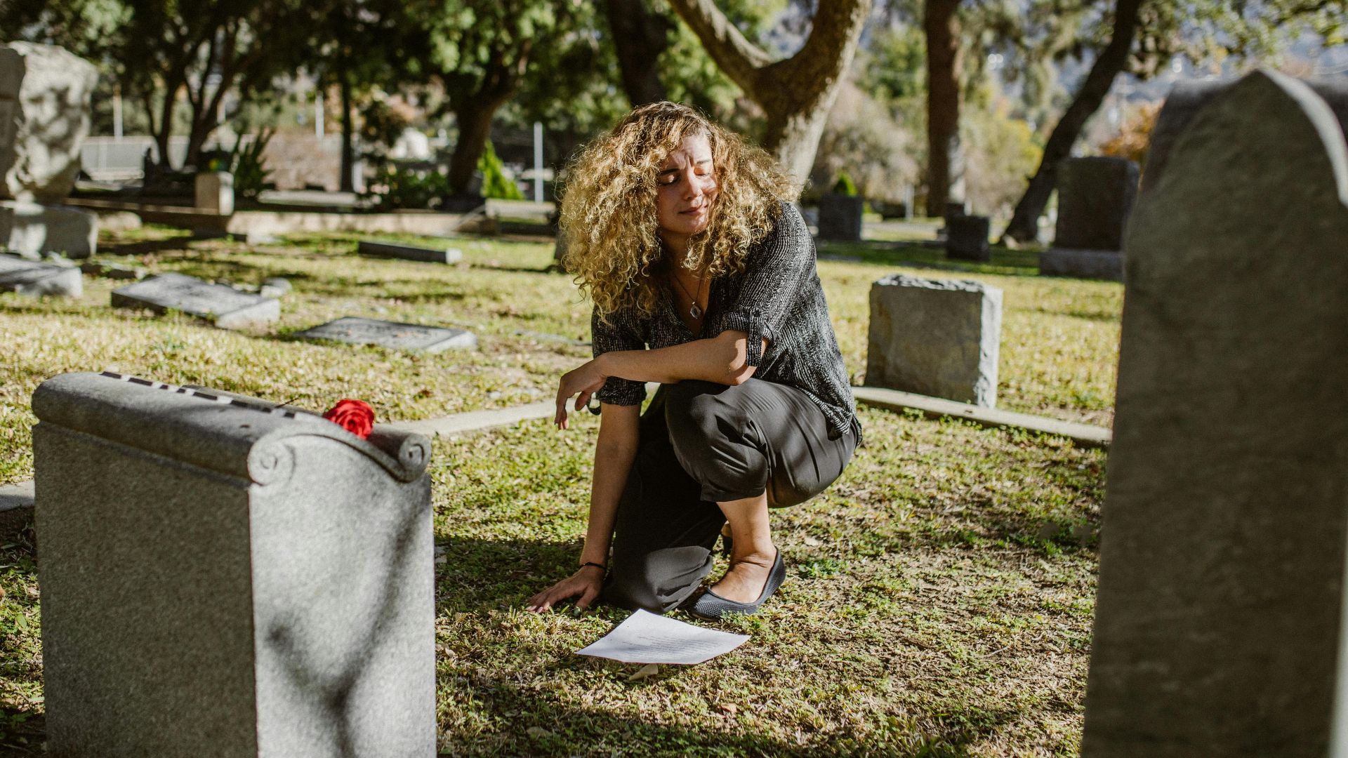 A woman kneeling by a grave in a sunny cemetery, expressing grief and bereavement.