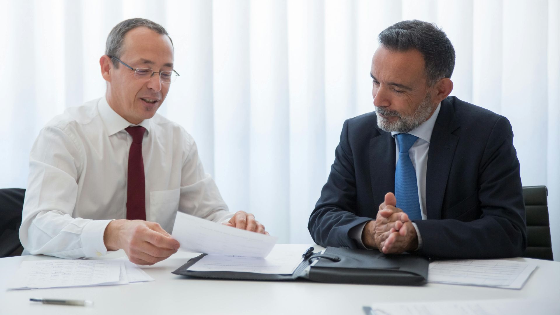 Two businessmen in suits having a discussion over documents in a bright office setting.