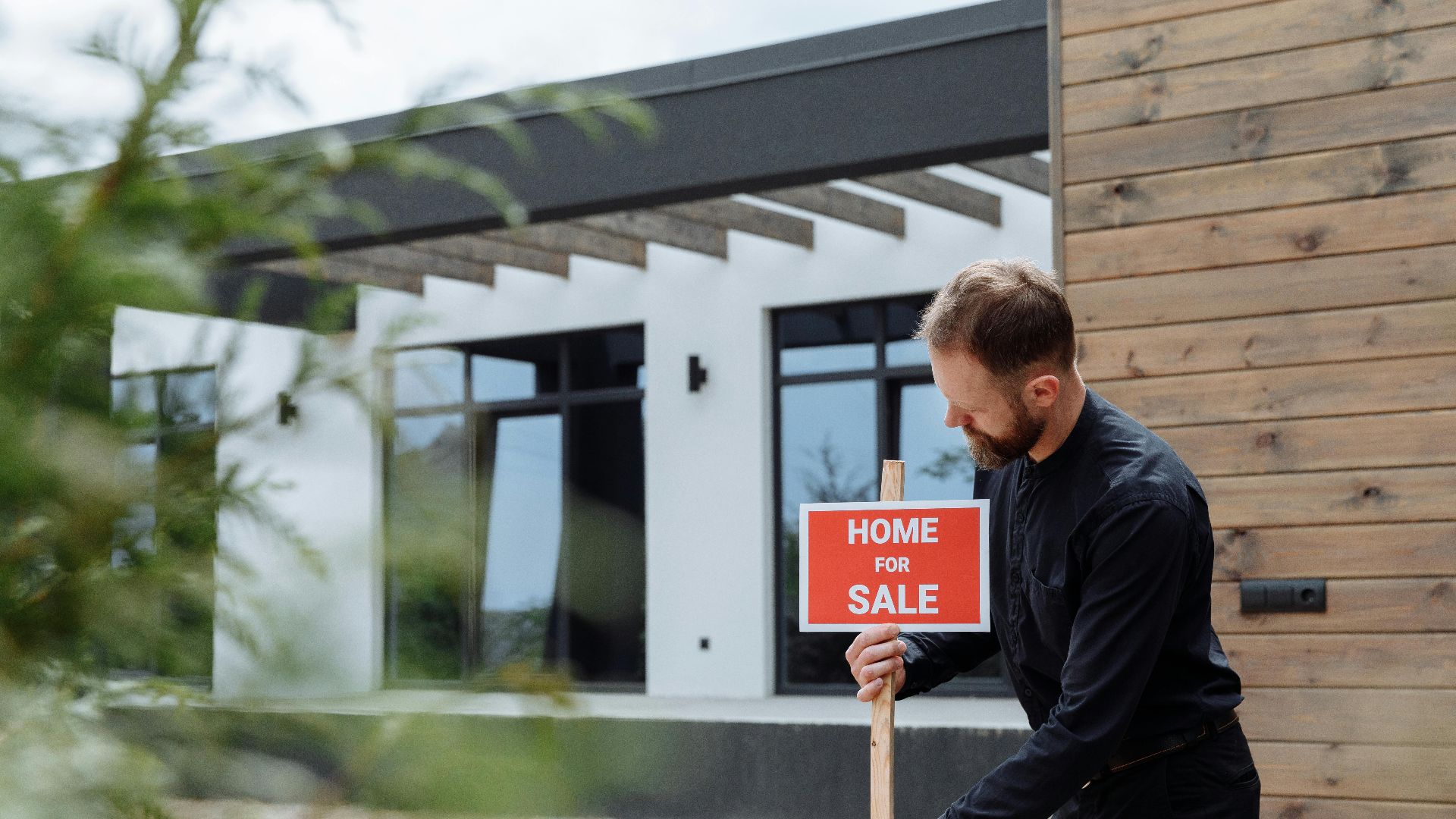 Man placing a home for sale sign outside a modern house for real estate listing.