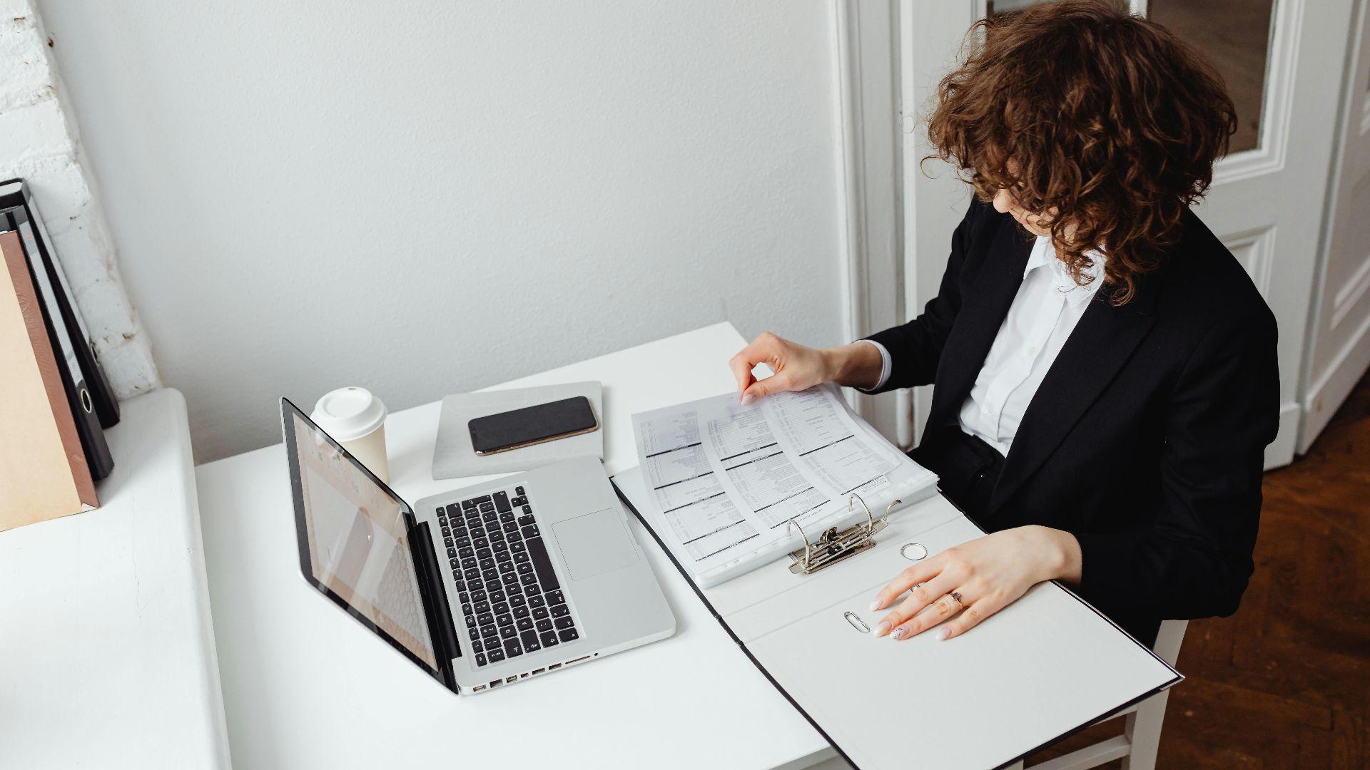 Professional woman in a home office reviewing documents on a laptop and papers.