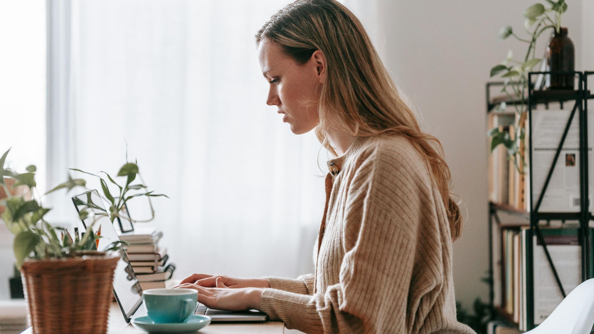 Side view of focused young female distance worker surfing internet on netbook at home desk with cup of tea
