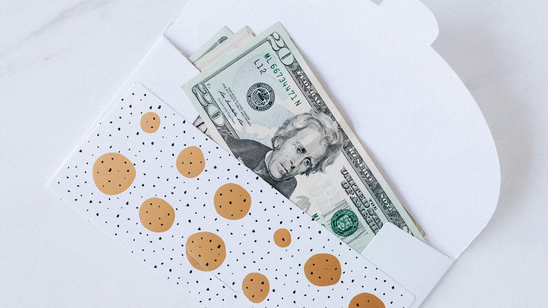 A polka dot envelope partially revealing US dollar bills on a white marble surface.