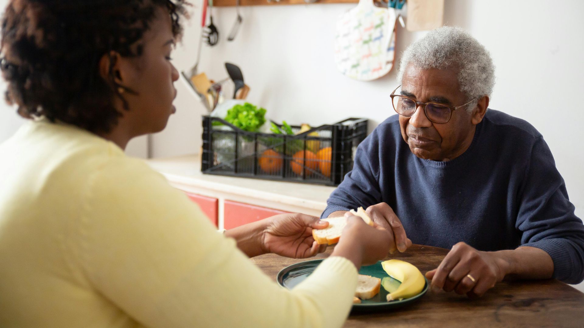 A woman assists an elderly man with meals at home, showcasing care and support.