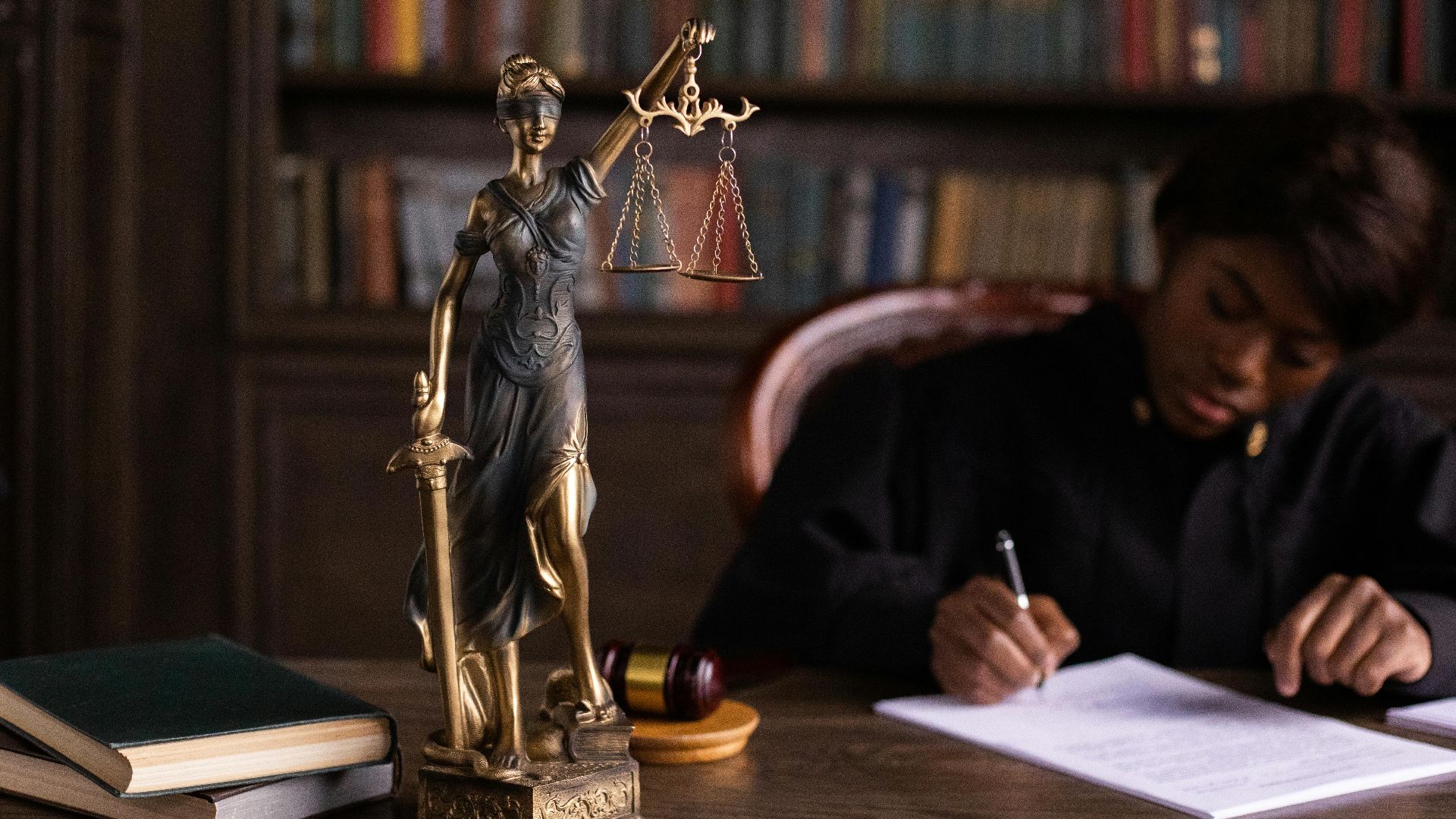 A focused judge writing on documents beside a Lady Justice statue in an office.