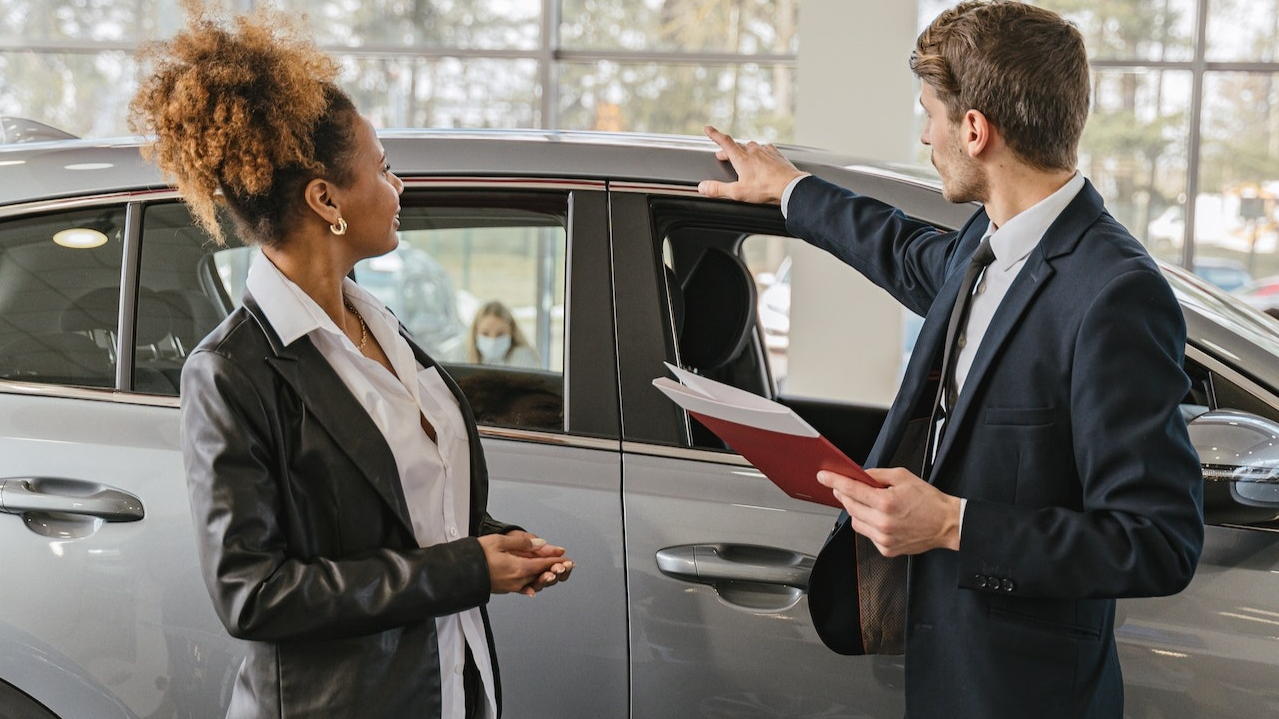 A woman is talking with a man wearing black suit in car dealership.