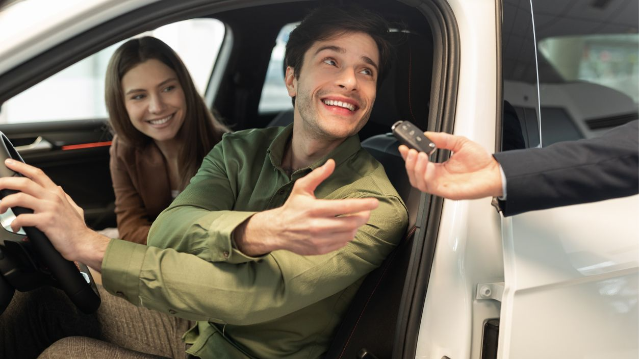 Happy millennial couple taking car key from auto salesman, sitting inside modern automobile at dealership, panorama. Cheery young family buying new vehicle at modern showroom