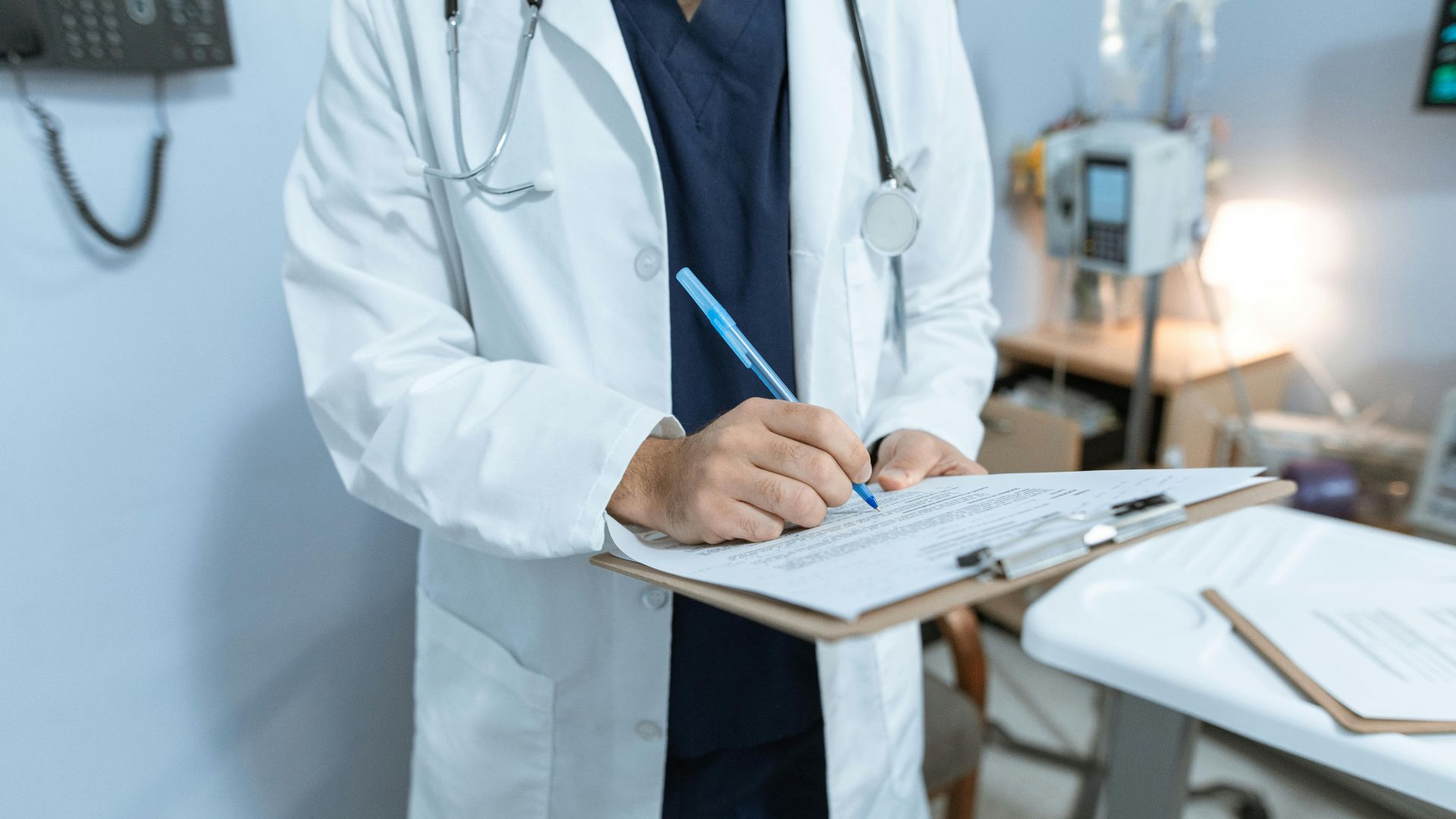 A doctor in a lab coat writing notes in a hospital setting, using a clipboard.