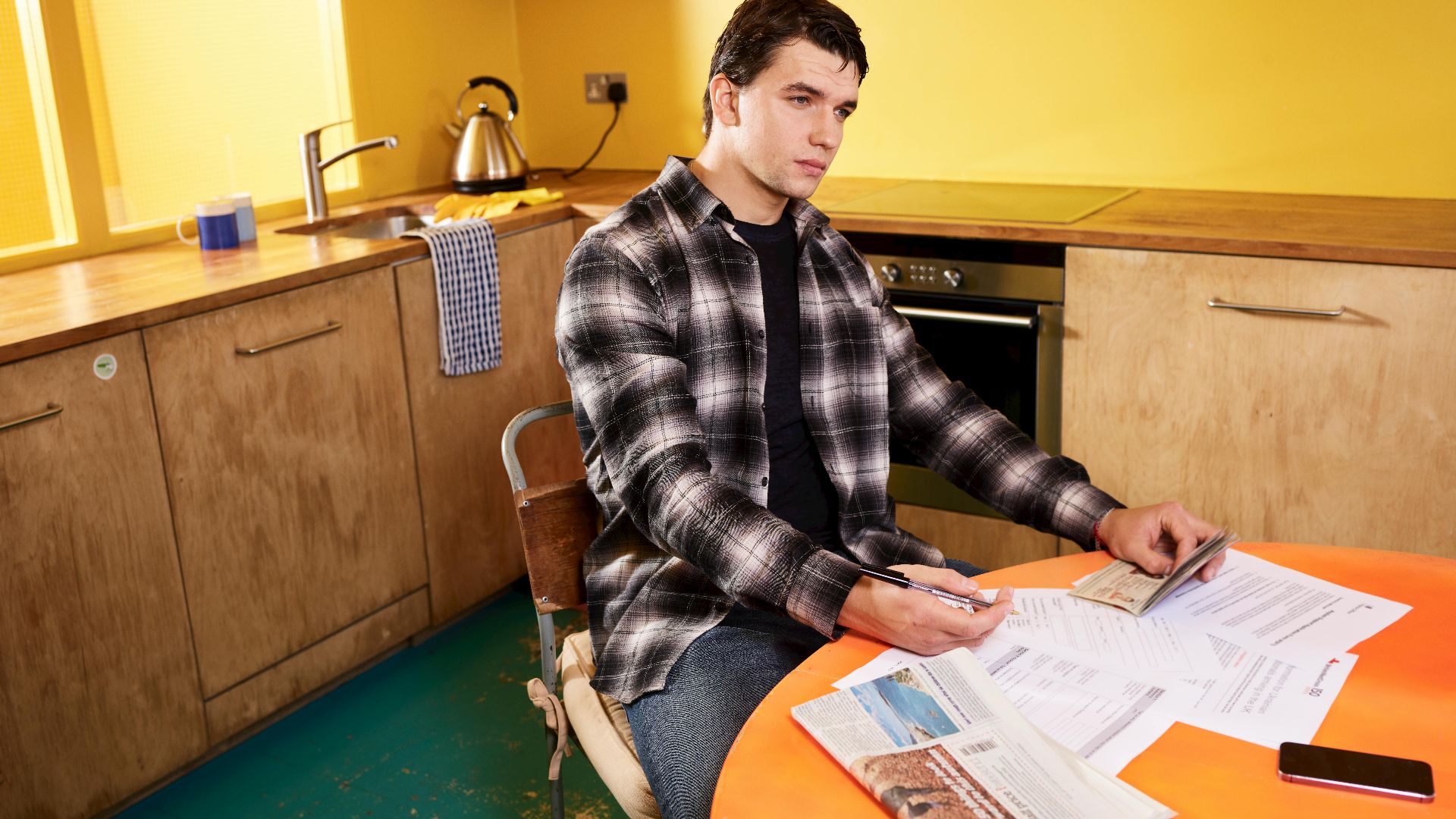 Man sitting in kitchen reviewing bills and a check with smartphone nearby.