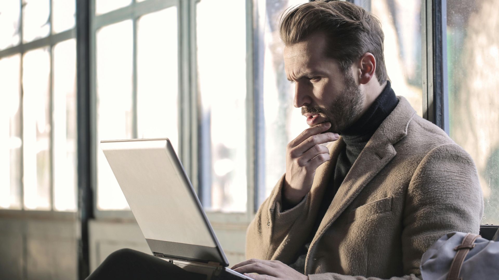 Man looking contemplative while working on a laptop in a well-lit indoor space.