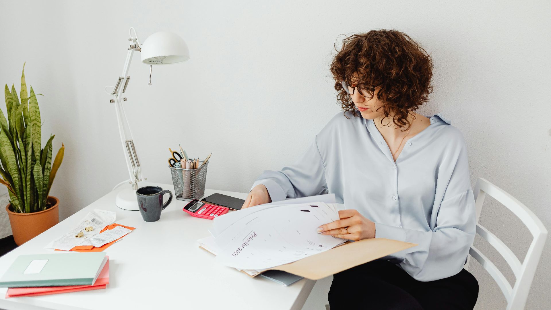 Woman reviewing documents in home office with plant and lamp.