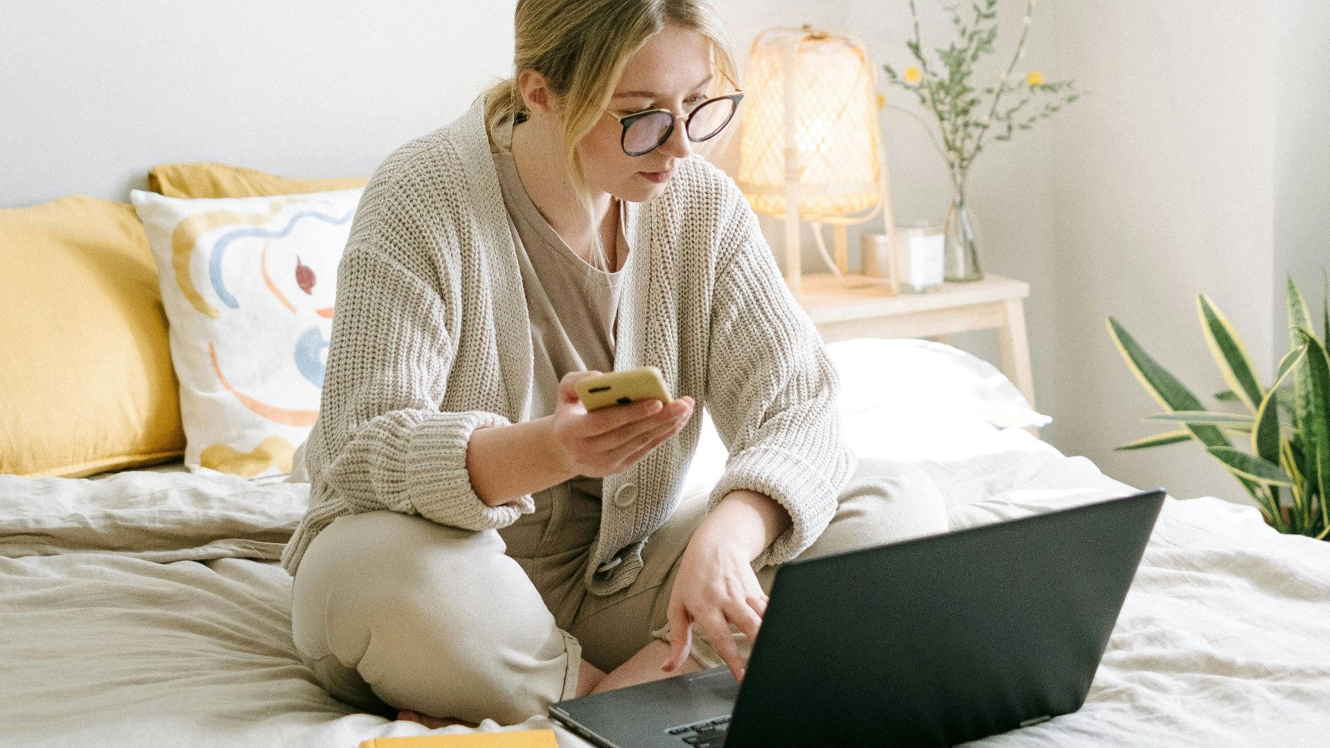 A woman working remotely from home sitting on a bed using a laptop and phone in a cozy bedroom.