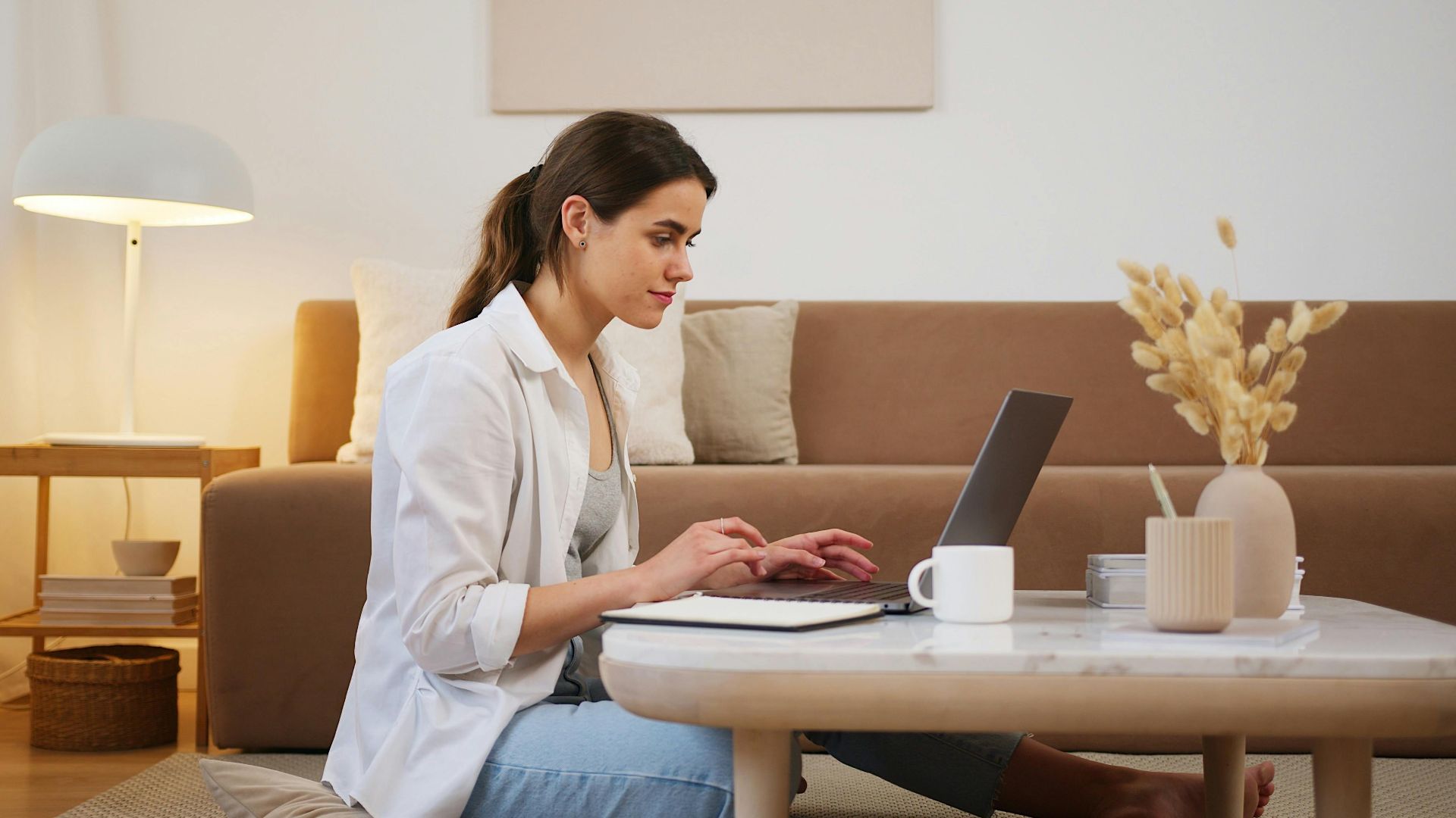 Side view of positive young lady with ponytail in casual outfit browsing contemporary netbook on coffee table while sitting on floor in cozy living room