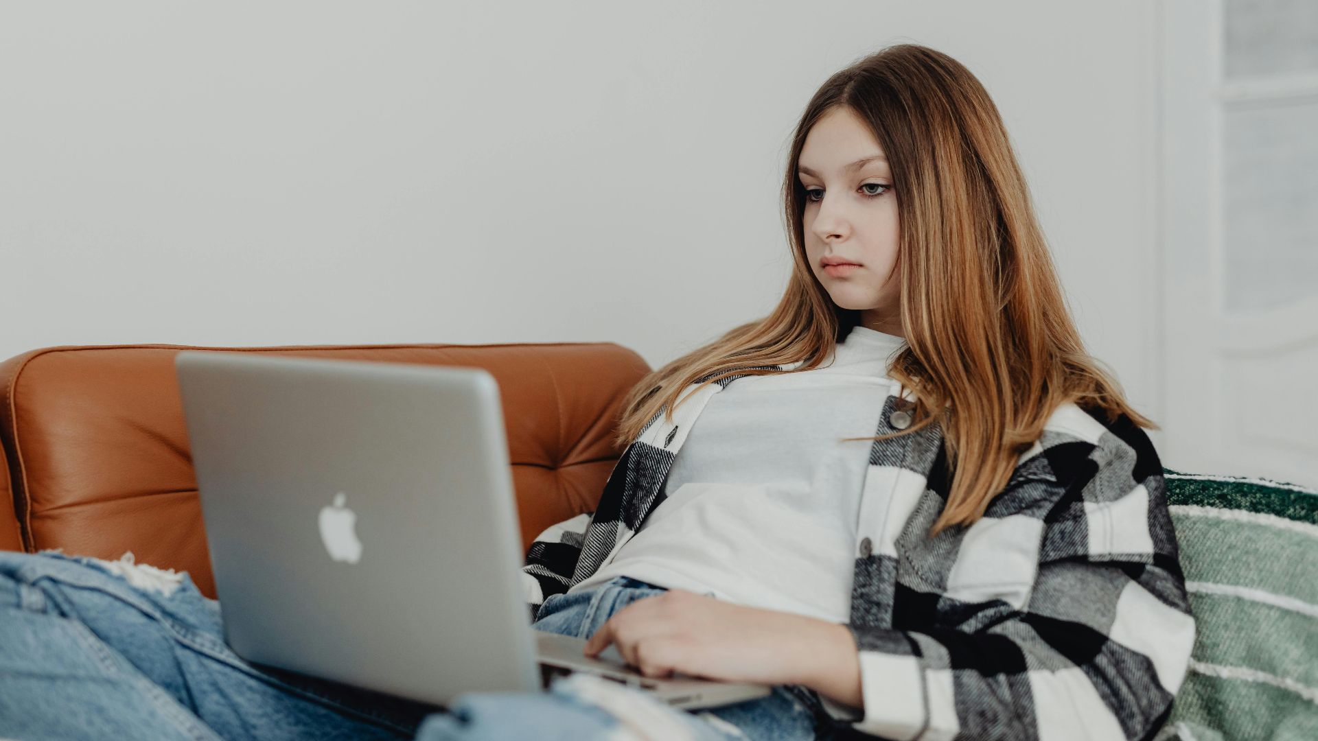 Teenage girl with long hair focused on laptop while lounging on a comfortable brown sofa indoors.