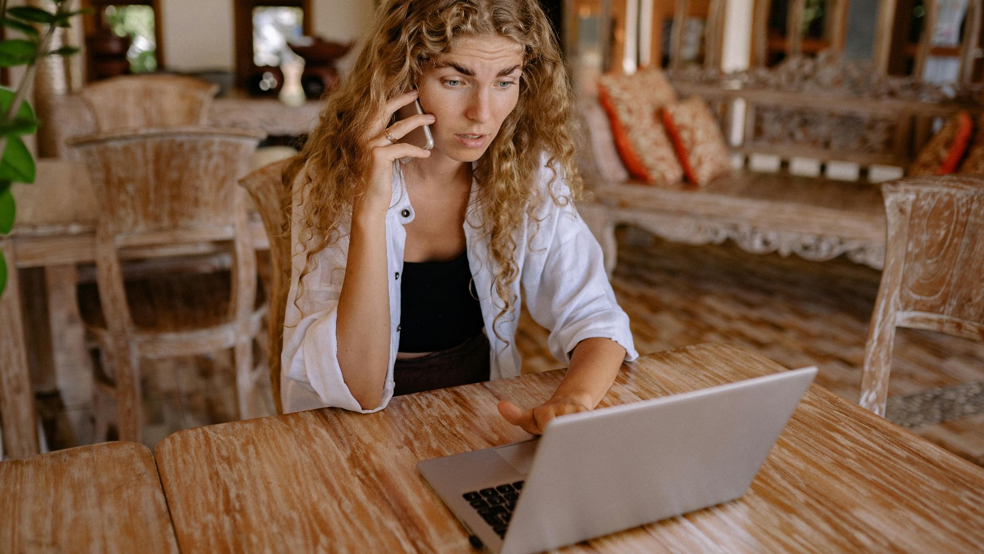 Woman sitting at wooden table, using laptop and talking on phone in a cozy indoor setting.