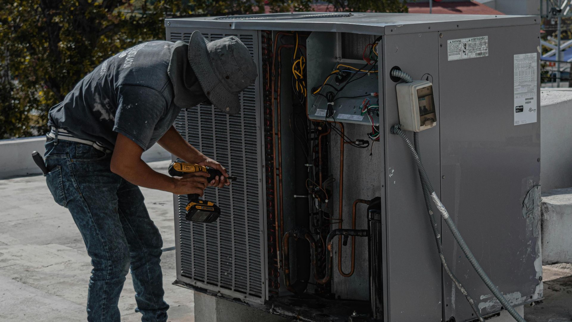 A worker in a bucket hat repairs an outdoor air conditioning unit on a rooftop.
