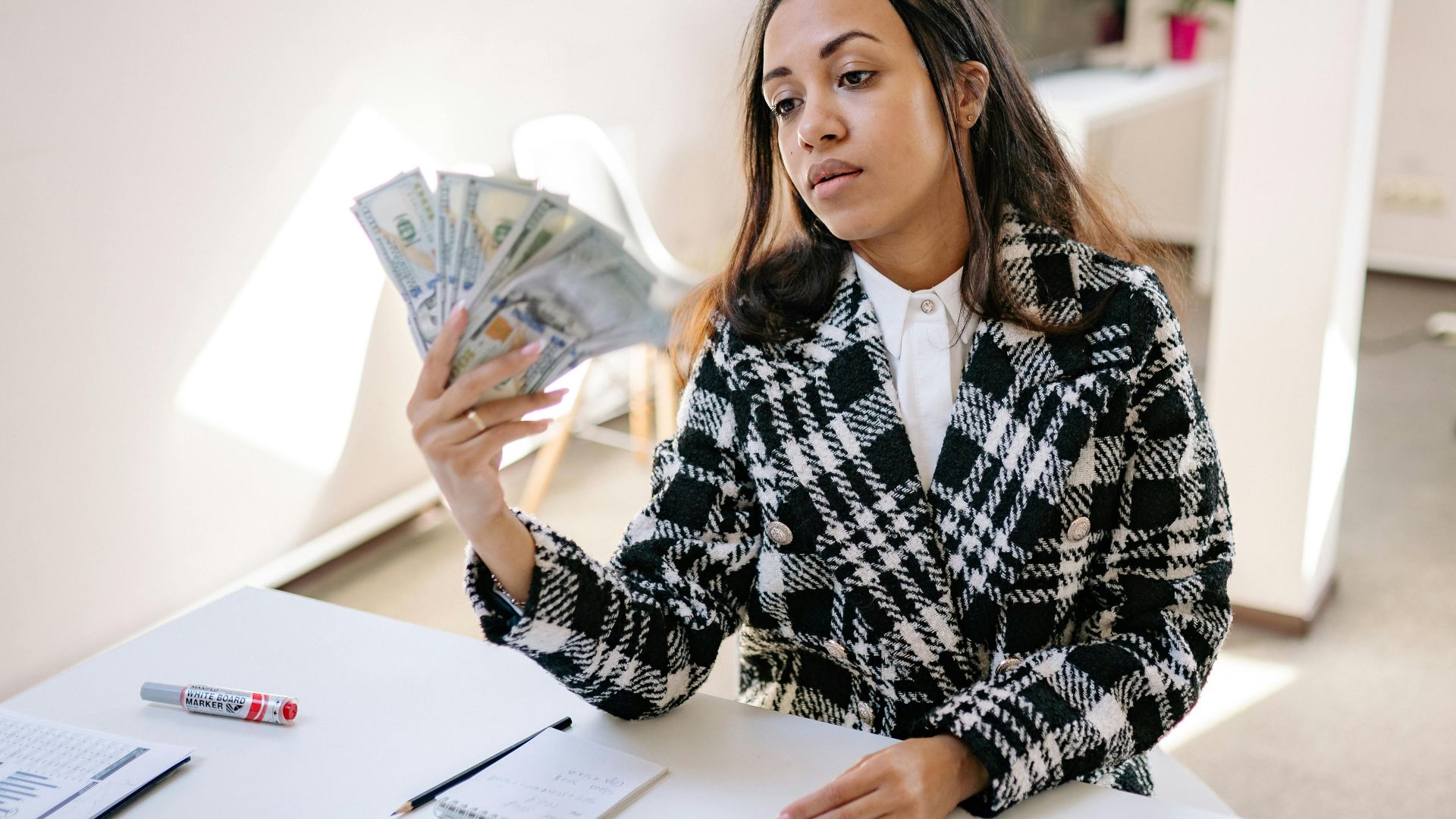 A young woman in a plaid coat counting US dollar bills at a desk in a modern office.