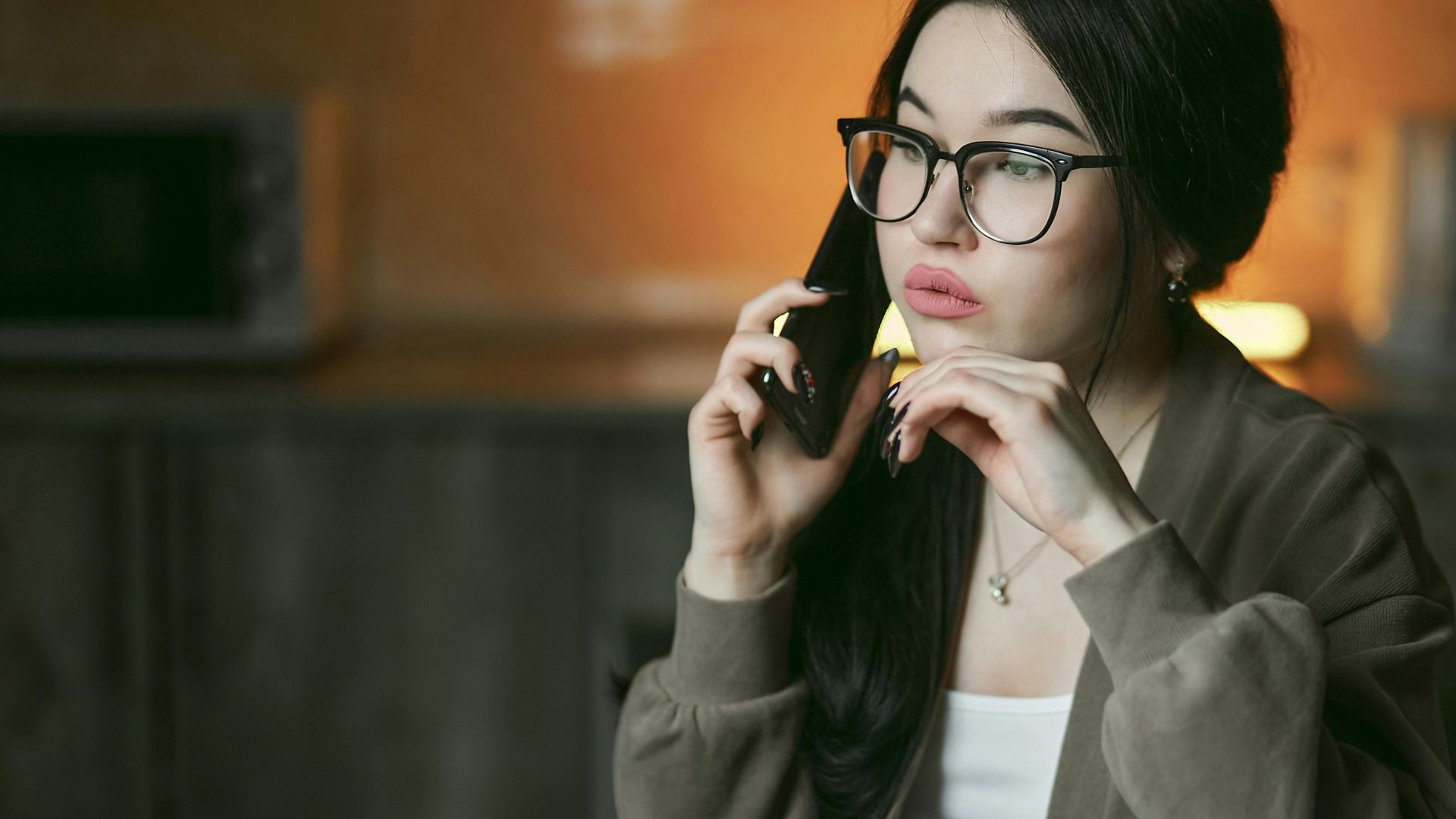 A woman with eyeglasses engaged in a phone call indoors, focused and thoughtful.