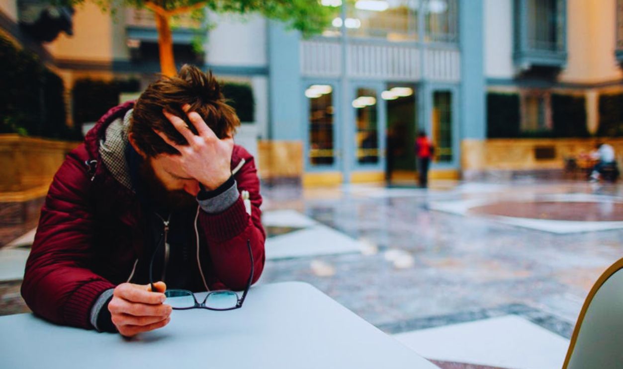 Selective Focus Photography of a Man Holding His Head and Eyeglasses Sitting Beside a Table