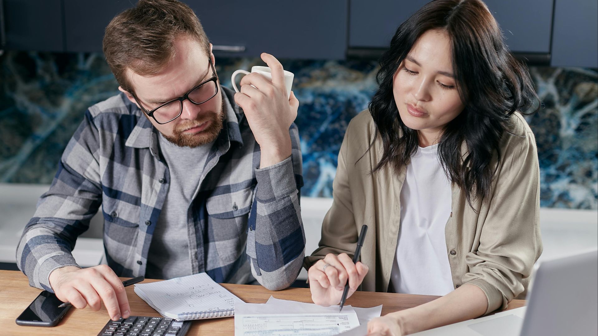 A couple reviewing household bills and budget using a calculator and laptop at their kitchen table.