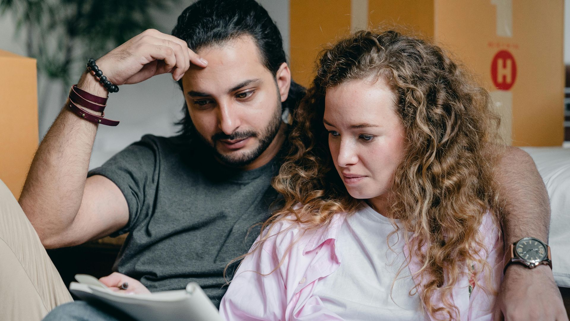 Focused male and female in casual wear sitting near boxes and looking through notebook thoughtfully while moving house
