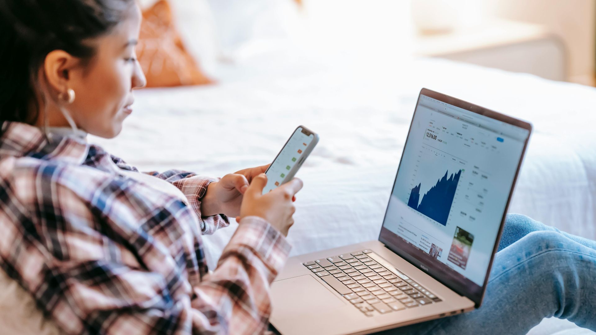 Side view of crop ethnic female dealer using trading app on cellphone against netbook with graph on screen in house