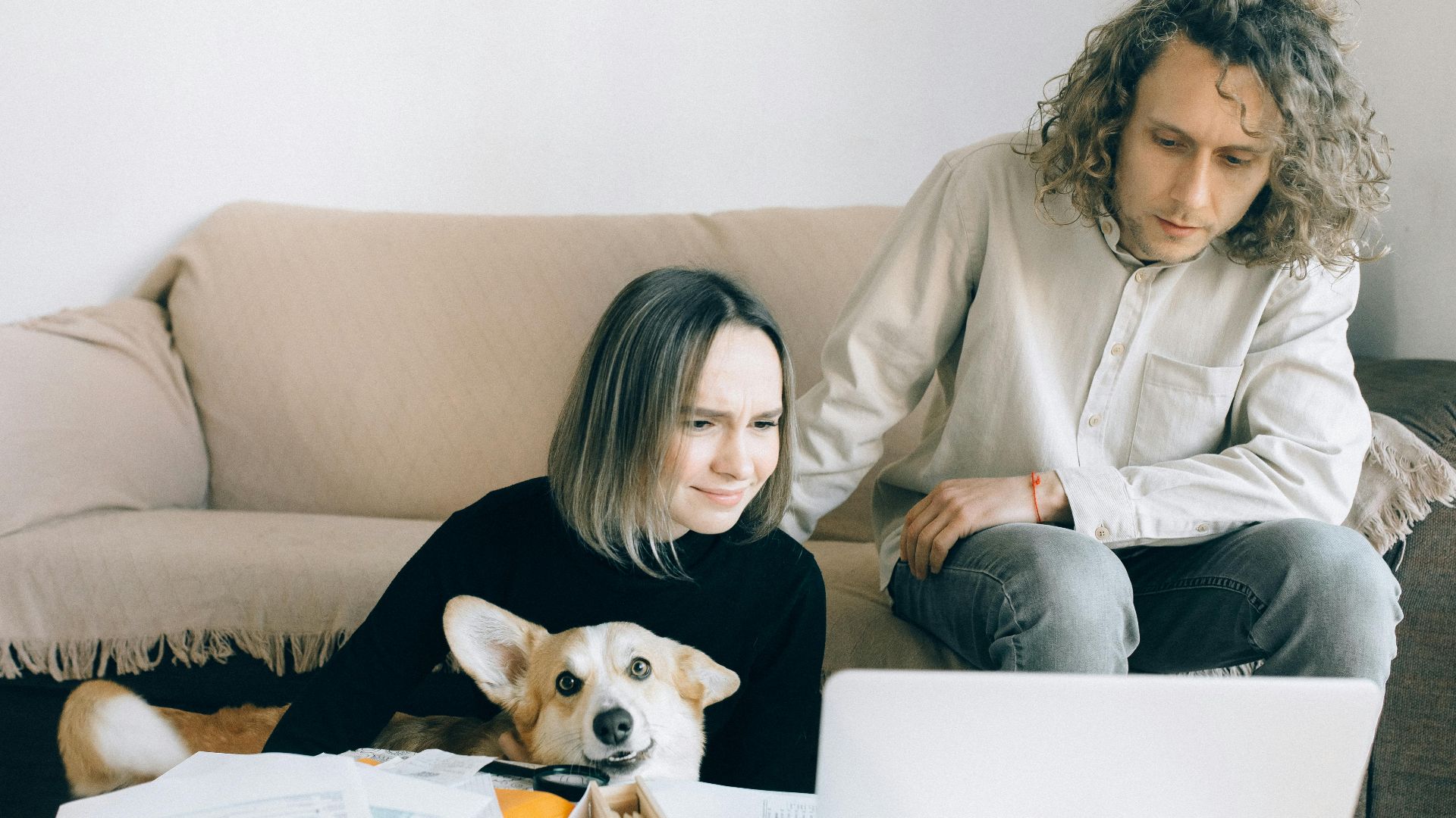 A couple looking at a laptop while sitting on a couch with their pet Corgi amidst paperwork indoors.