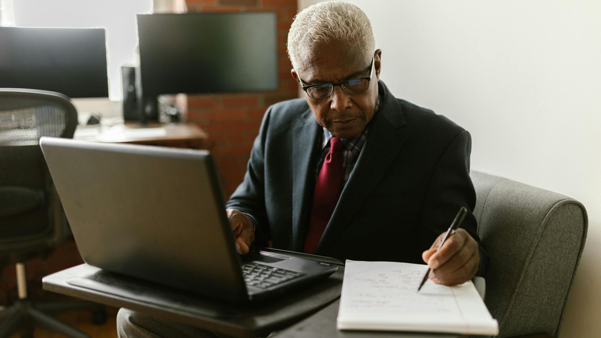 Elderly businessman in a suit writing notes while using a laptop in a modern office setting.