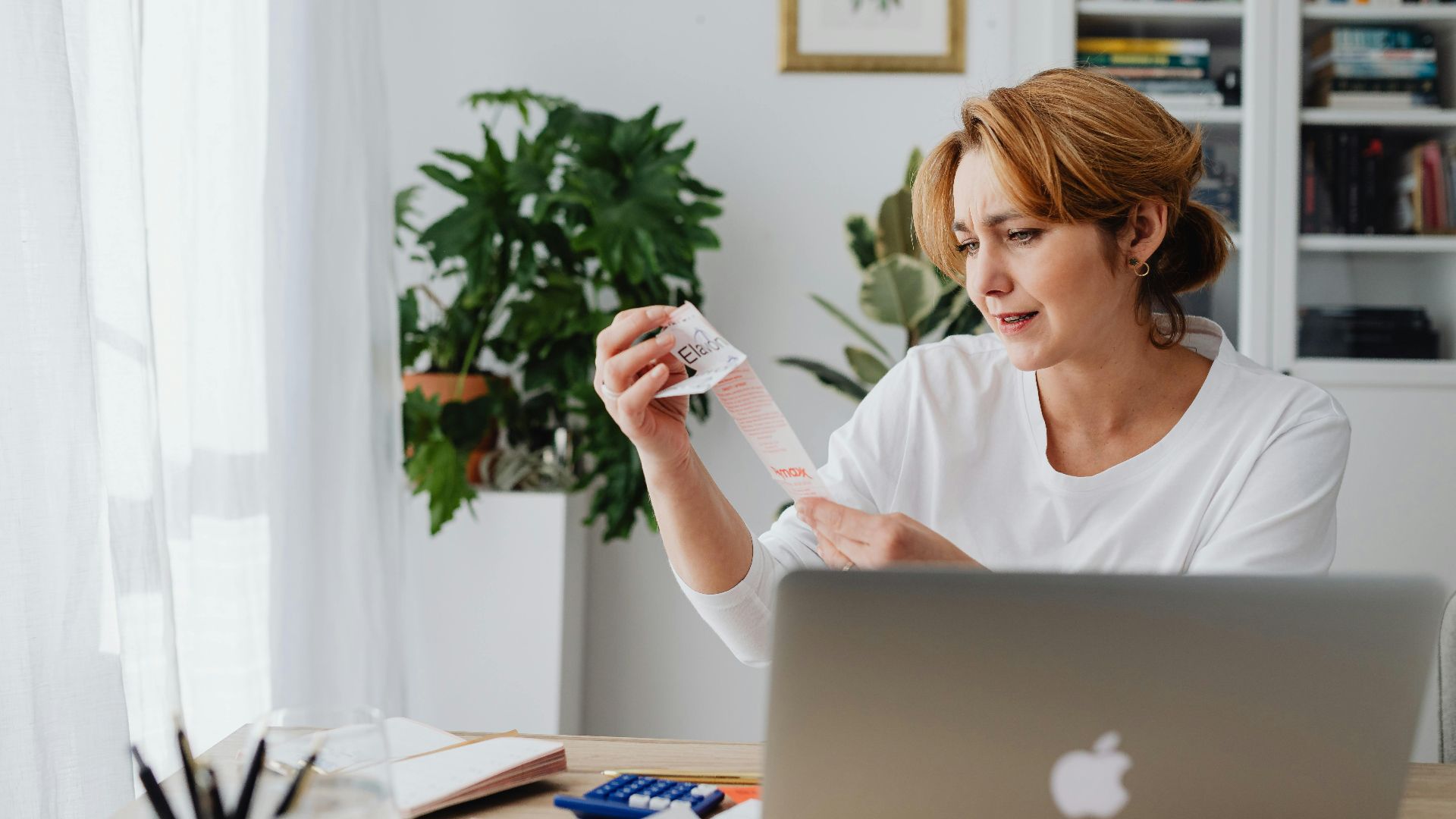 A woman in a white shirt sits indoors, examining a lengthy receipt with a concerned expression.