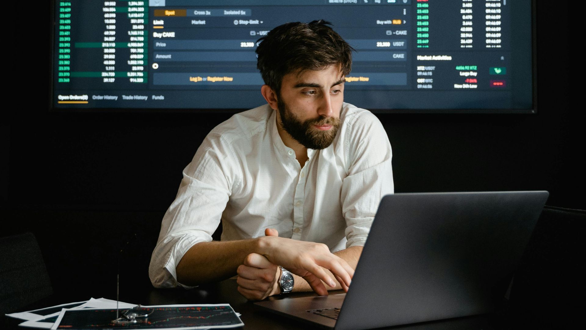 A man analyzing stock market data on a laptop in an office setting with a projector screen displaying financial charts.