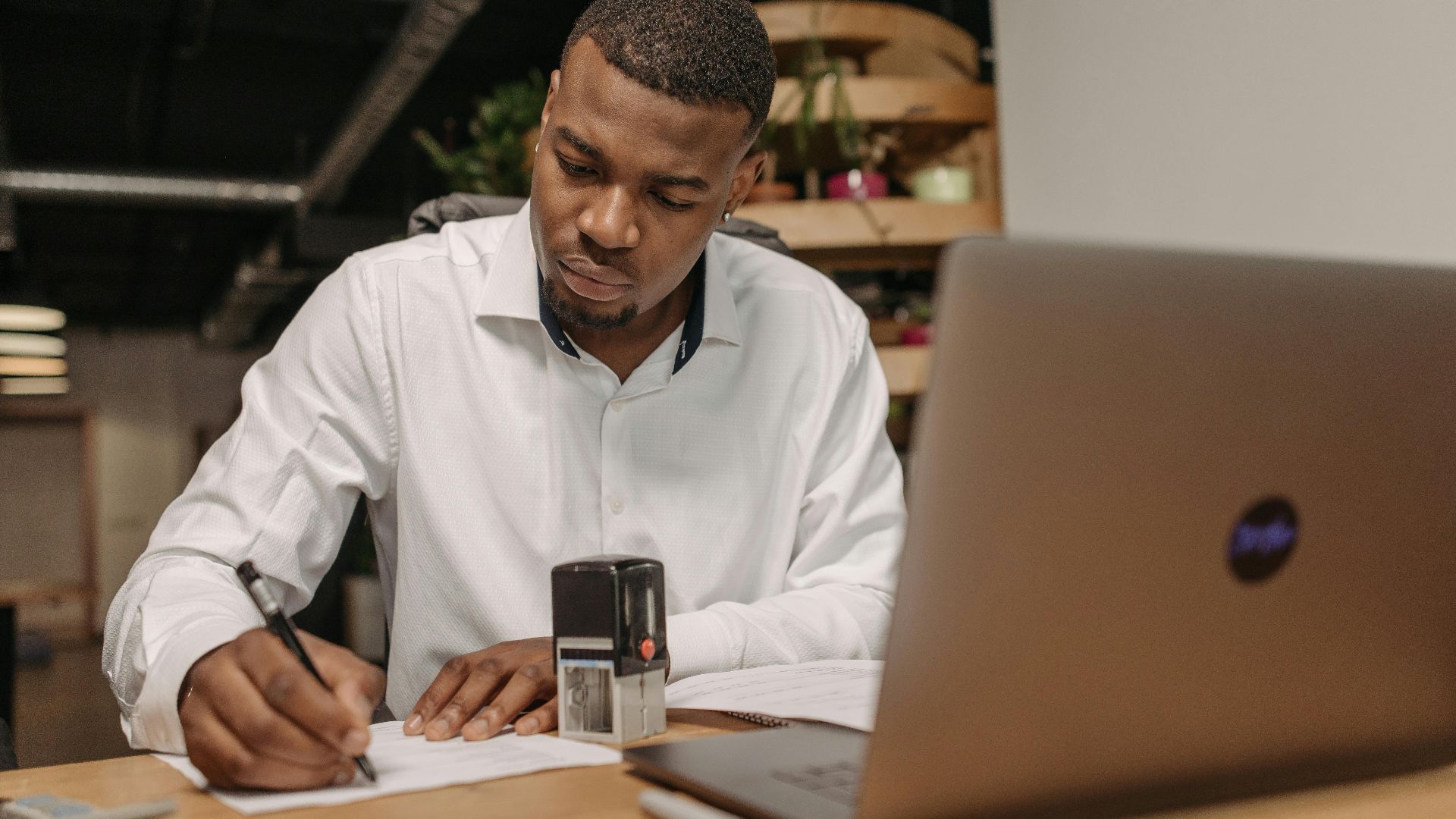 A young man in a white shirt works diligently at his office desk with a laptop and documents.