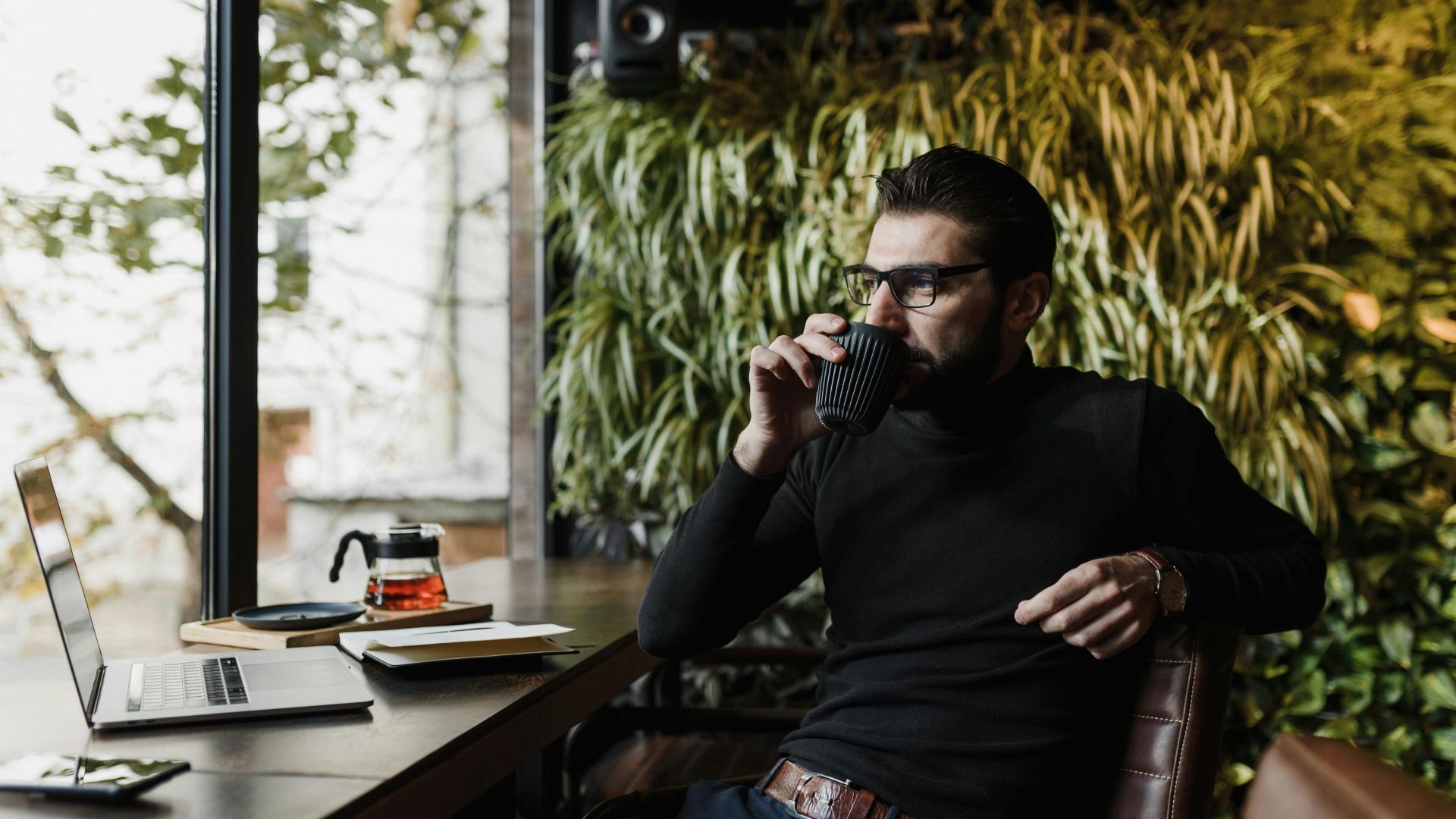 Young man enjoying a coffee while working on a laptop in a cozy cafe with lush greenery.
