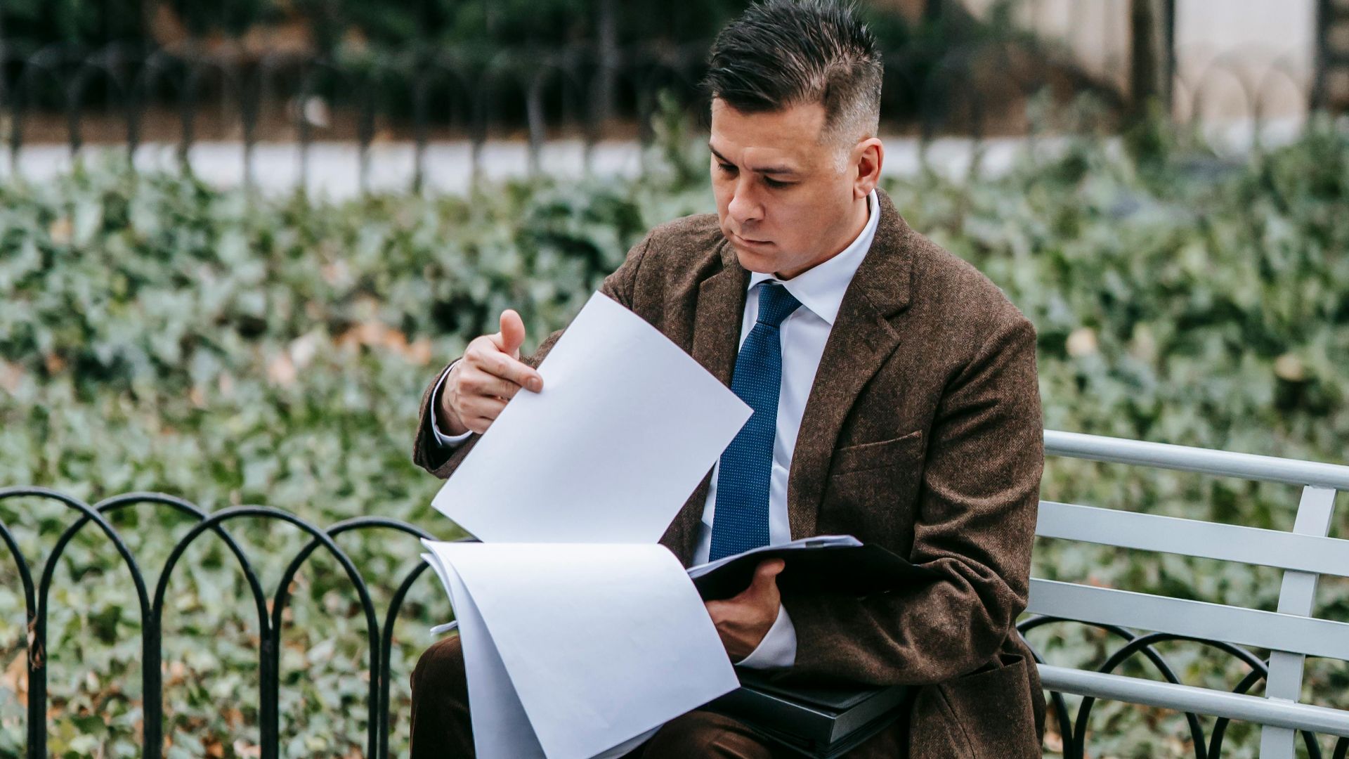 Professional man in formal wear reviewing documents while sitting on a park bench outdoors.