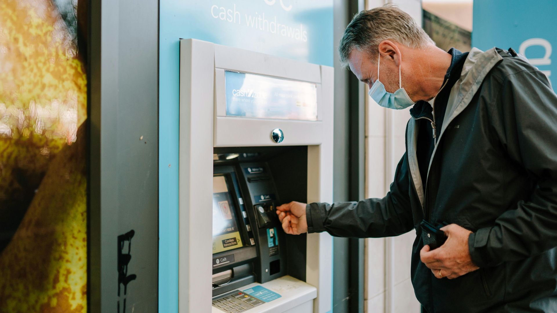 A man wearing a mask using an ATM machine outdoors for cash withdrawal.