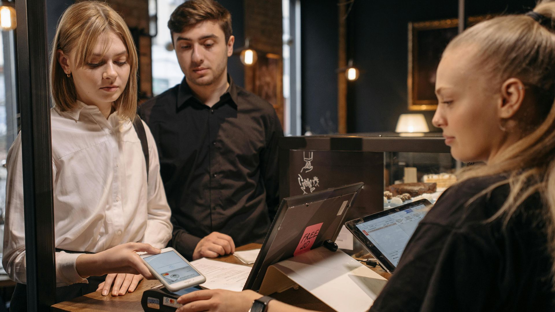 Two customers making a contactless payment at a café counter using a mobile phone.