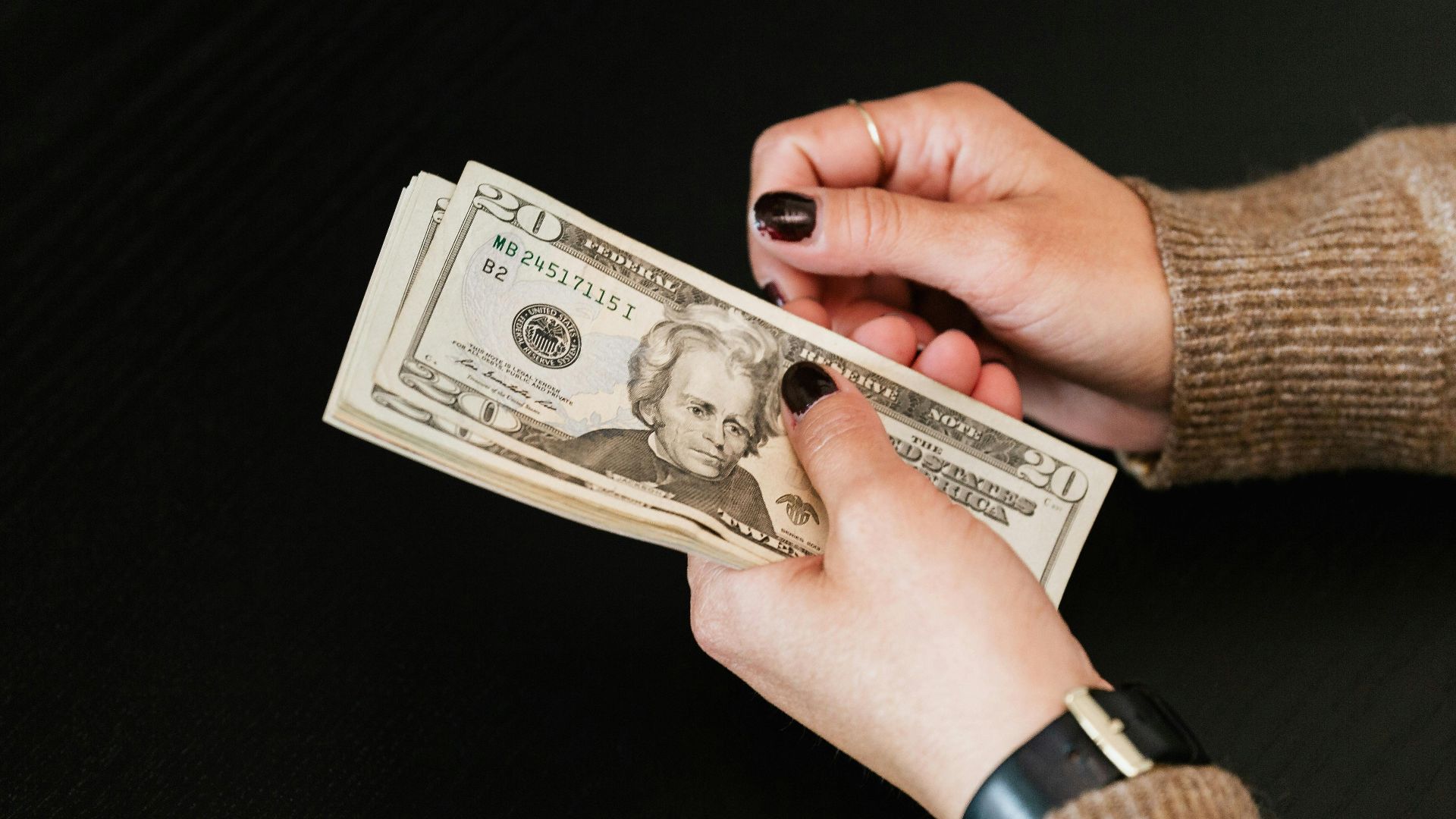 Close-up of hands counting a stack of US dollar banknotes on a dark table.