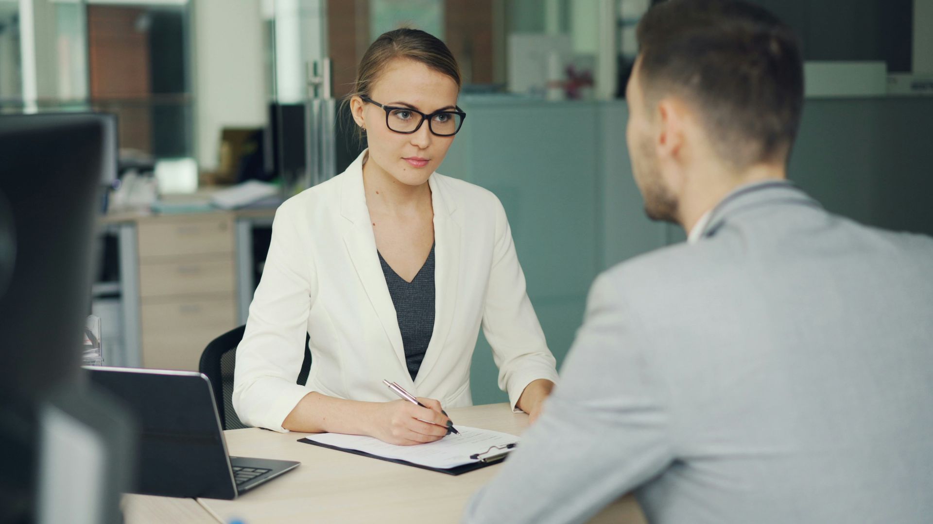 Woman in glasses interviews man at office desk.