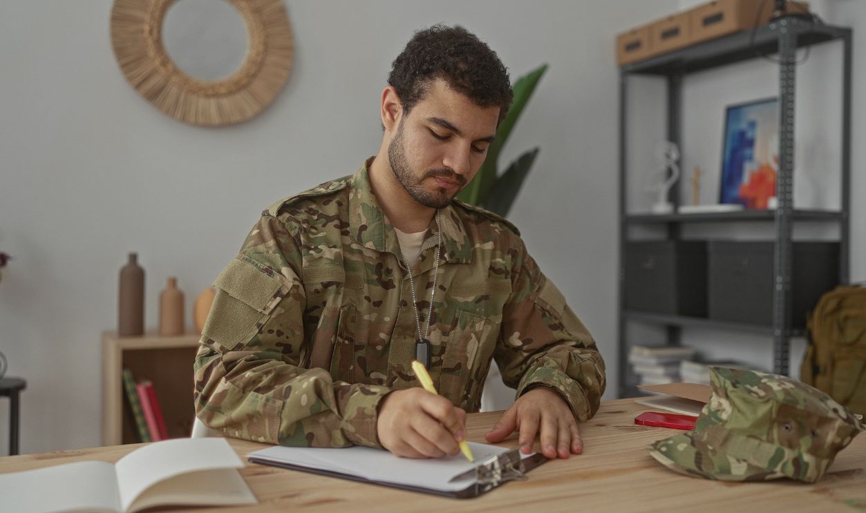 Man writing on clipboard with right hand at desk in military building interior wearing camouflage uniform and dog tags; duty.
