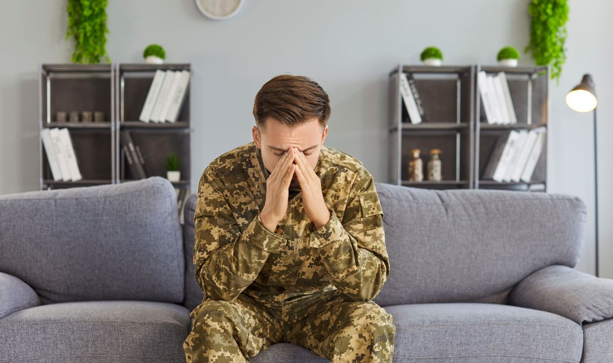 Sad man soldier sits on sofa at home, facing depression and mental health stress, dealing with post trauma, reflecting personal struggle in a quiet domestic space with emotional weight.