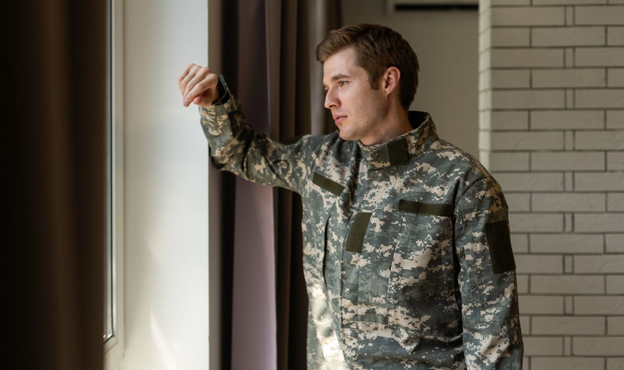 Depressed and sad soldier in green uniform with trauma after war standing near the window