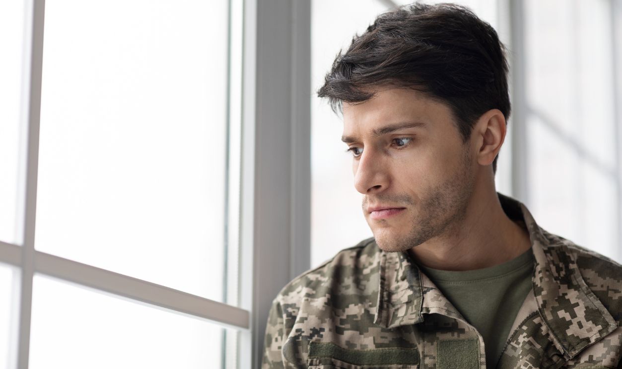Serious short-haired young man in military uniform soldier looking through the window, upset soldier suffering from posttraumatic syndrome after returning home, copy space, closeup