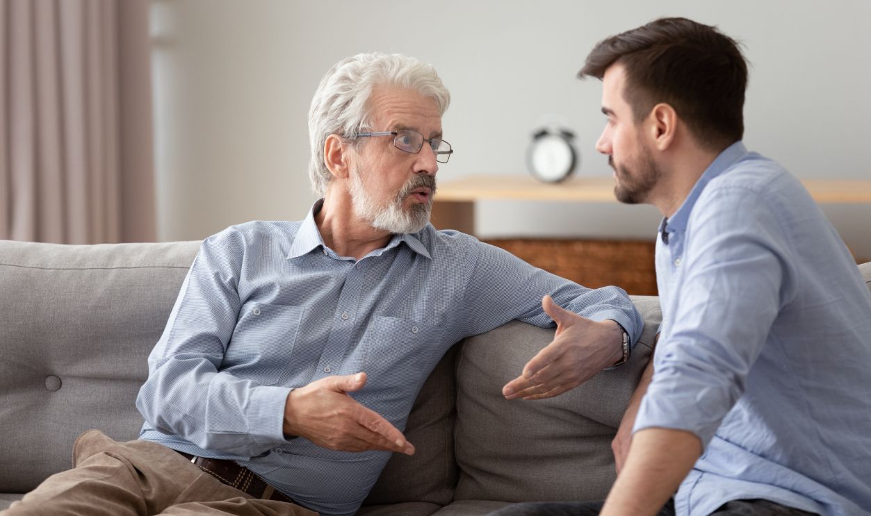 Serious 60s elderly father and grown up adult son sitting on sofa talking having important conversation trying to solve life issues problem, different men relative people communication at home concept