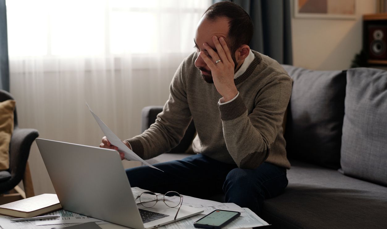 Young troubled man touching his head and looking through financial bill while sitting on couch in front of table with laptop and papers