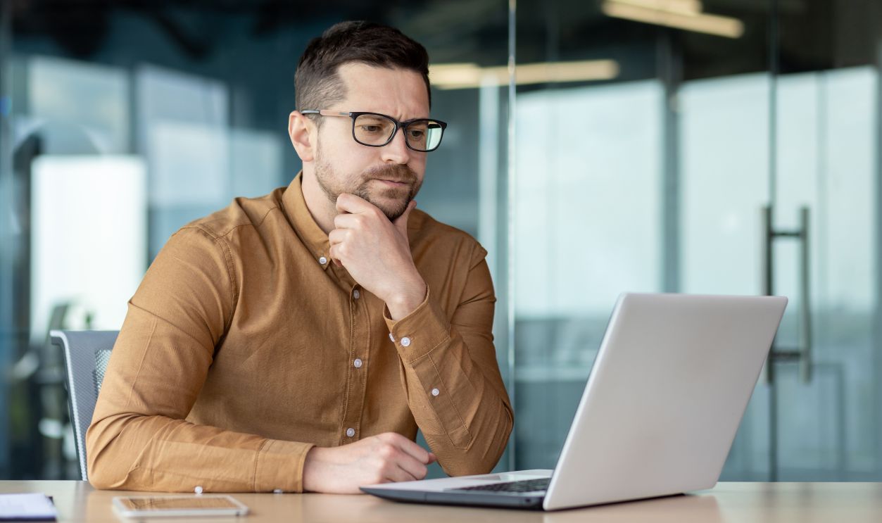 Confused young male businessman sits in the office in front of a laptop and looks thoughtfully at the screen, concentrates on a new project, solves a problem.