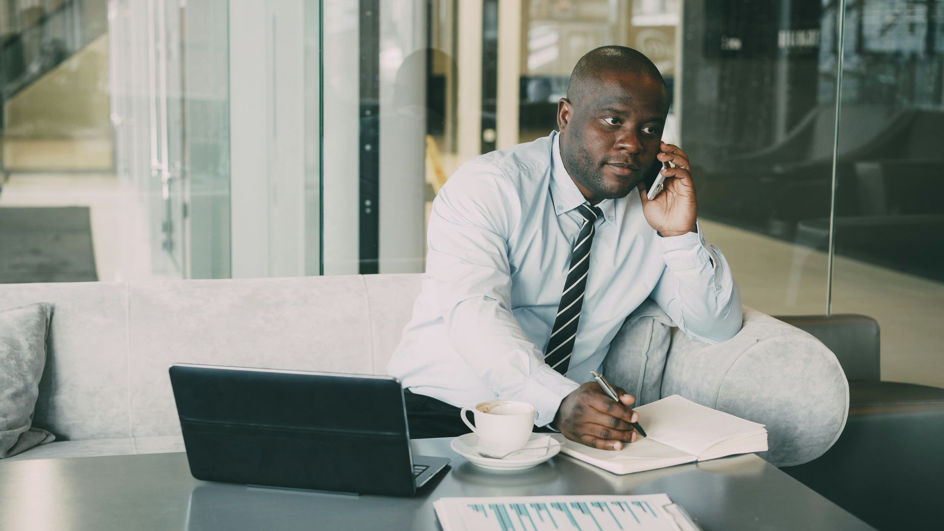 Man on phone, writing in notebook at desk.
