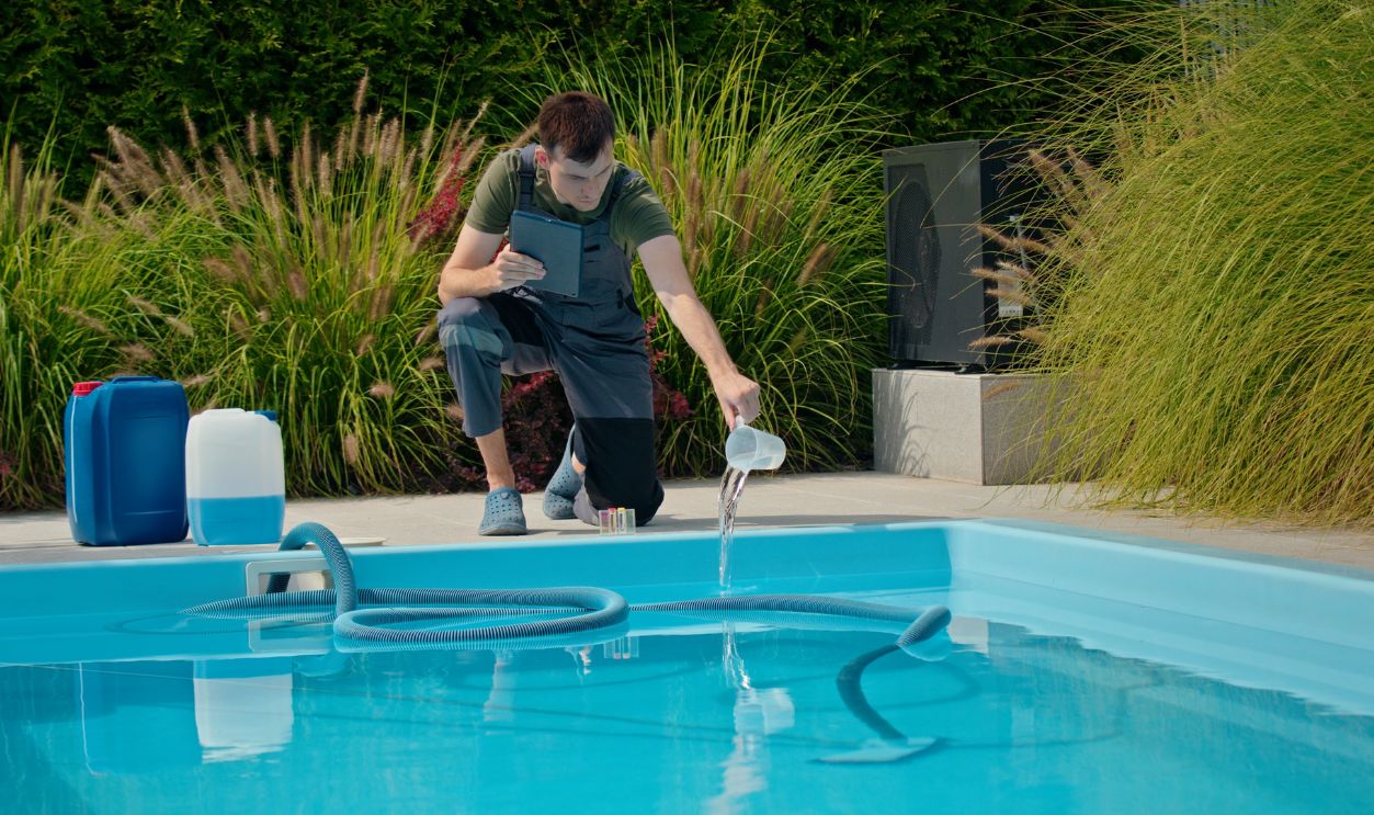 Pool technician pouring liquid into the pool while using a tablet, with chemical containers and greenery in the background.