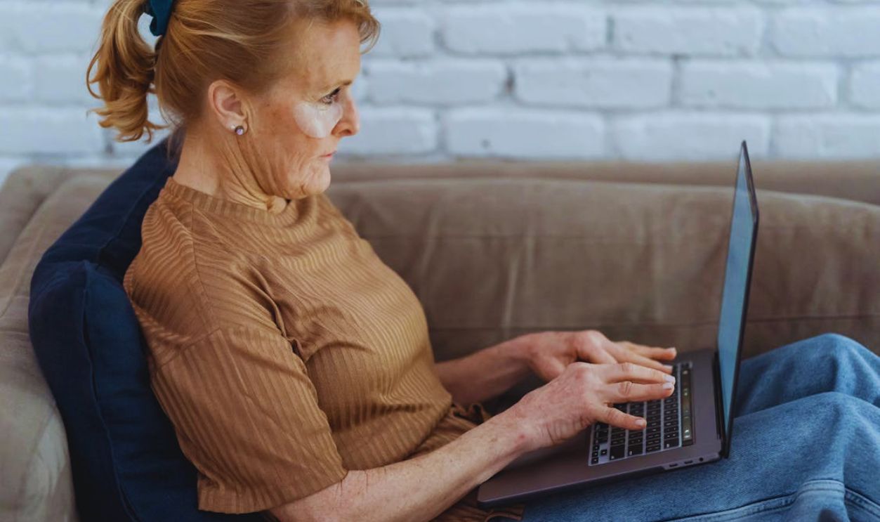 Crop mature woman working on laptop on cozy sofa