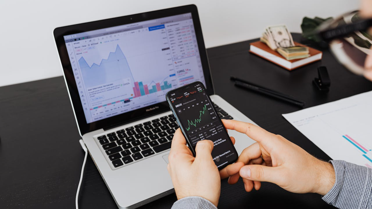 The image shows a close-up of a person's hands holding a smartphone over a laptop on a dark desk
